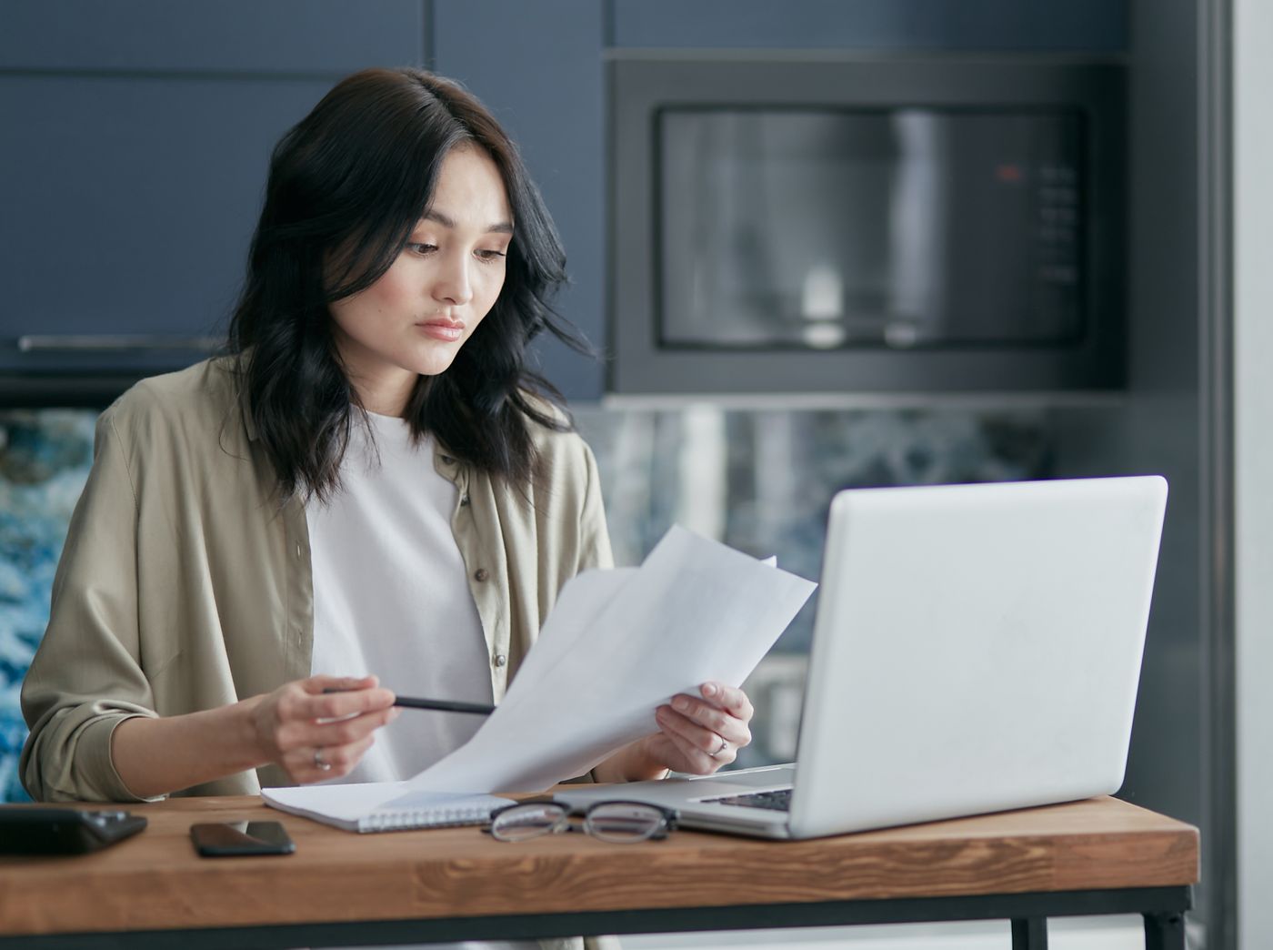 A woman at a home table going through paperwork with a laptop open.