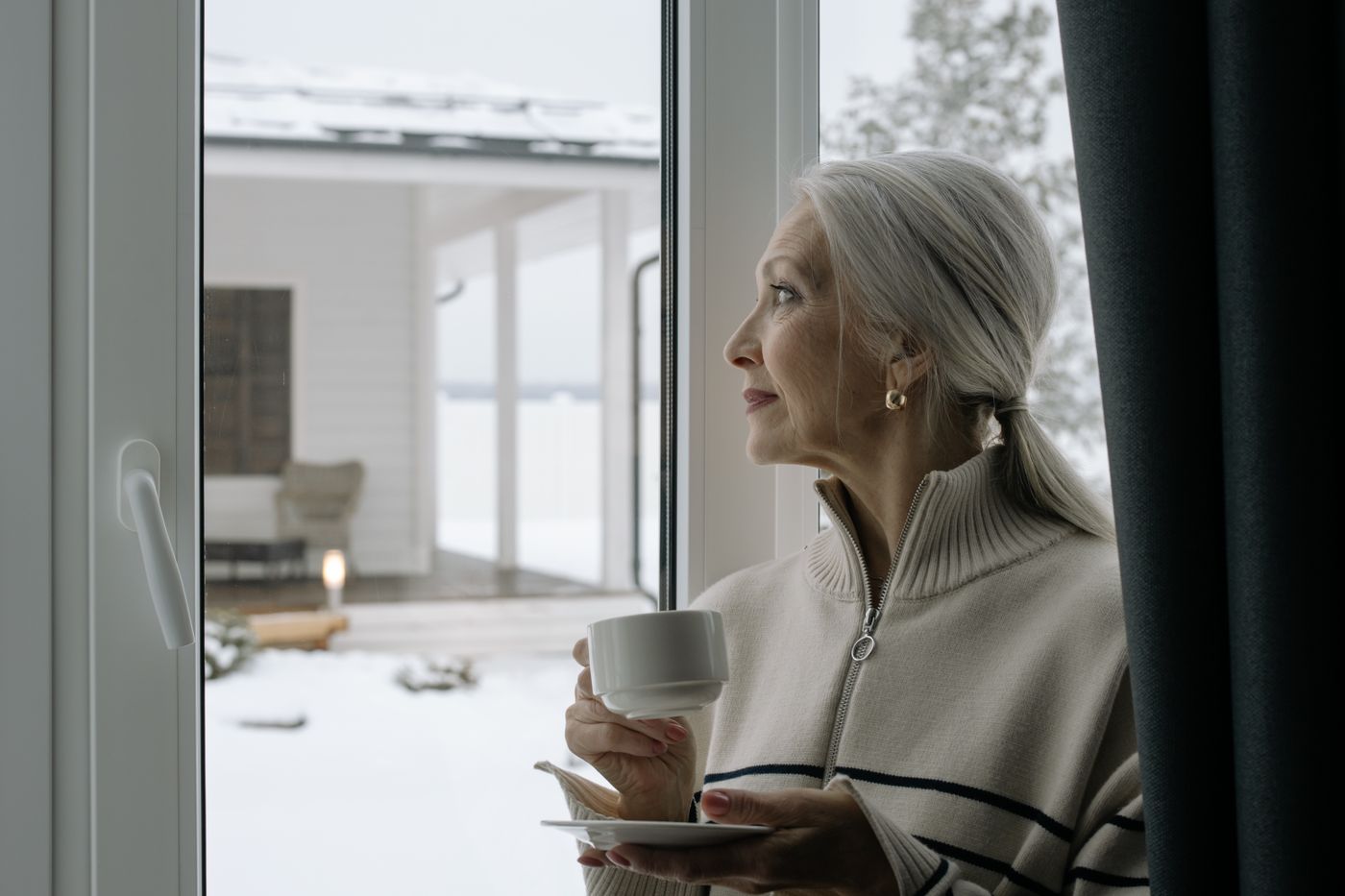 A woman with gray hair holds a cup of coffee and looks out a bright window, thinking.