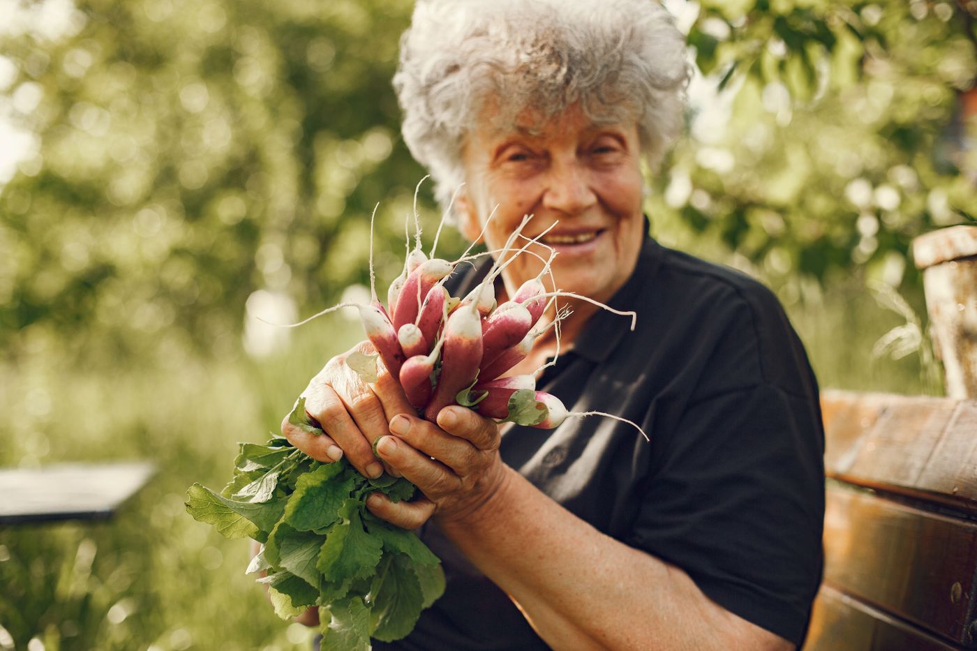 A smiling woman in her garden holding freshly picked vegetables.